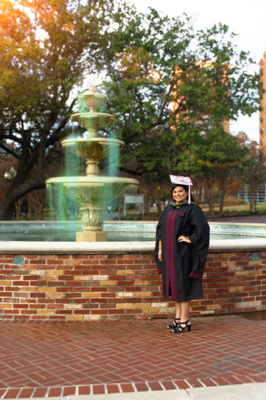Celebratory graduate in black cap and gown standing beside a brick fountain with sunlight filtering through green trees in the background.
